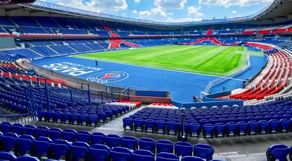 Vue panoramique du **Parc des Princes** vide, avec ses tribunes bleues et rouges, son terrain de football verdoyant et le logo du PSG au centre.