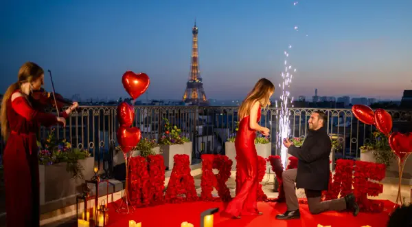 Proposition romantique à Paris, avec la Tour Eiffel en arrière-plan. Un homme s'agenouille, une bague à la main, devant sa compagne. Des roses rouges forment le mot "MARRY ME". Une violoniste joue, des feux d'artifice illuminent la scène. Un moment dig...