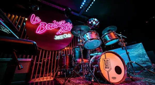 Batterie de jazz dans un bar parisien, éclairé par des néons roses et bleus. Ambiance feutrée pour une soirée musicale.