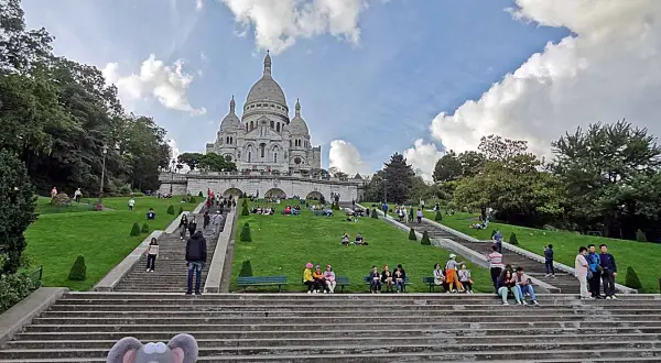 Un adorable raton laveur en pull rouge pose sur les marches menant au Sacré-Cœur, où des visiteurs profitent du soleil sur l'herbe verdoyante.