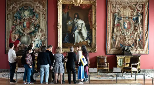 Visiteurs admirant le portrait de Louis XIV et des tapisseries grandioses, une scène digne des 50 plus beaux tableaux du Louvre.