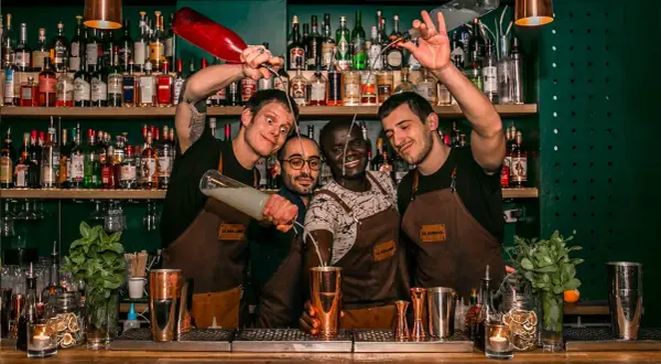 Quatre barmans joyeux préparent des cocktails au bar "Le Renard". L'un verse un liquide d'une bouteille rouge, un autre d'une bouteille transparente.