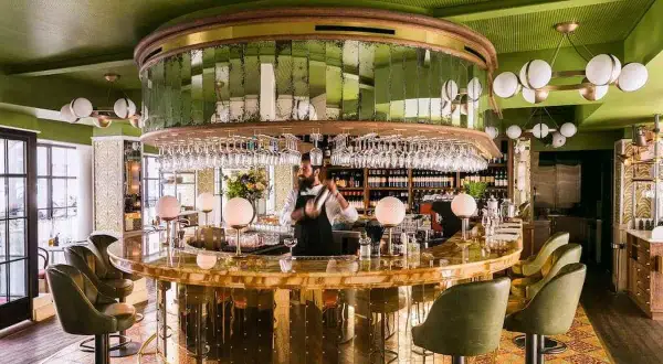Barman préparant un cocktail dans un bar chic aux murs verts et au comptoir doré. Une ambiance de "pink mama" pour une soirée réussie.