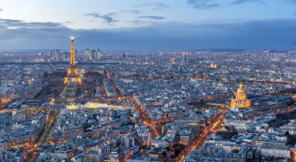Vue panoramique de Paris au crépuscule, avec la Tour Eiffel illuminée et le dôme des Invalides. On aperçoit le quartier de La Défense au loin. Un panorama depuis l'**étage tour Montparnasse**.