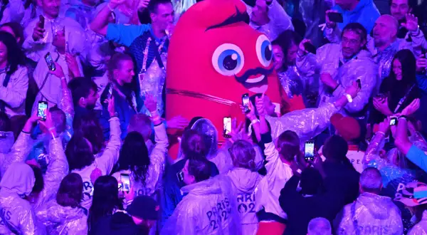 La Phryge JO, mascotte des JO, interagit avec une foule joyeuse. Les gens filment et prennent des photos avec leurs téléphones.