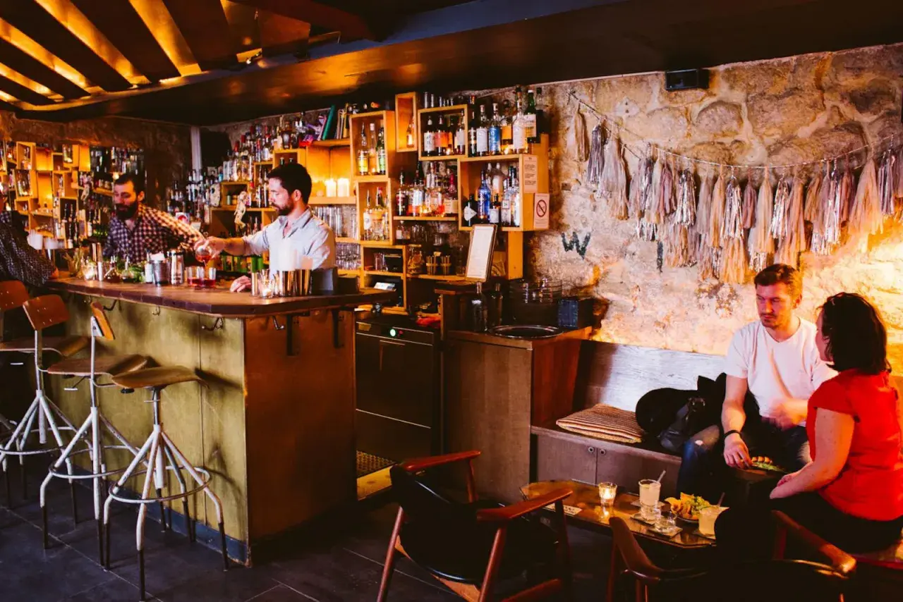 Ambiance feutrée dans ce bar caché. Un barman sert un cocktail, des clients discutent au comptoir et sur une banquette.