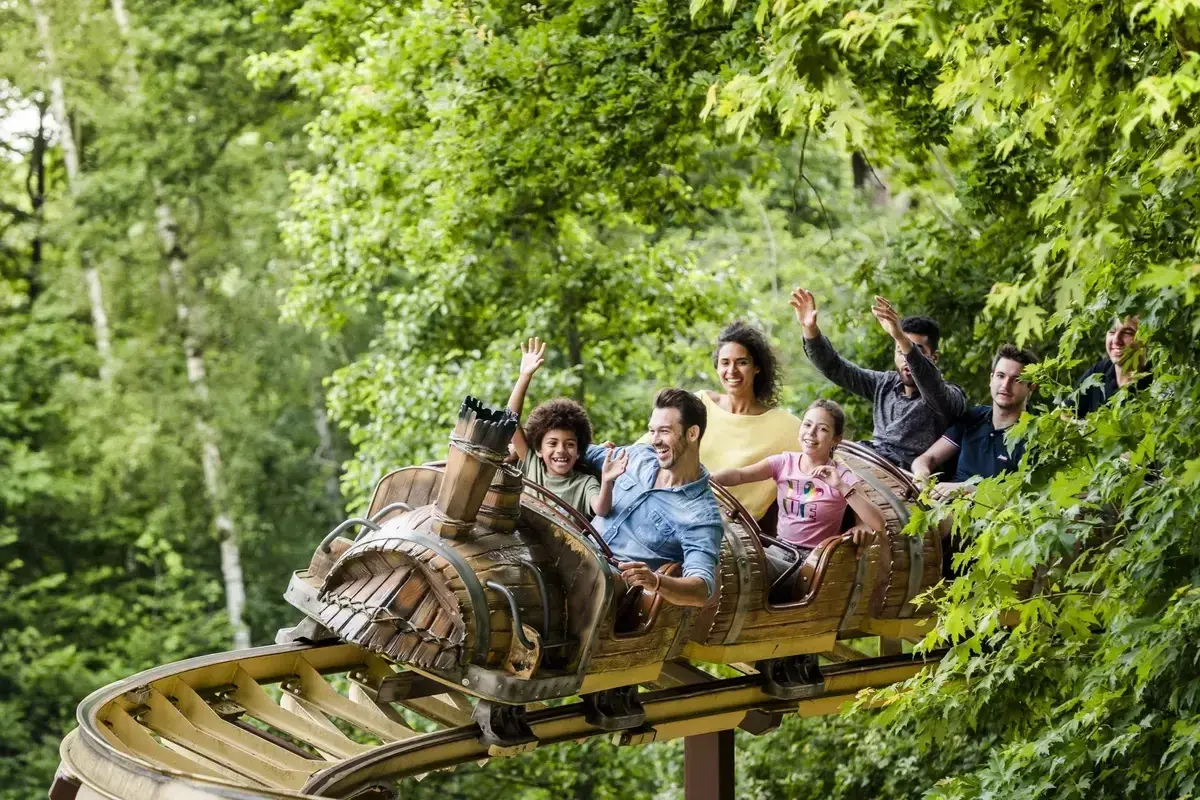 Famille souriante et joyeuse sur un wagon de parc attraction, bras levés, profitant d'une descente dans un décor verdoyant.