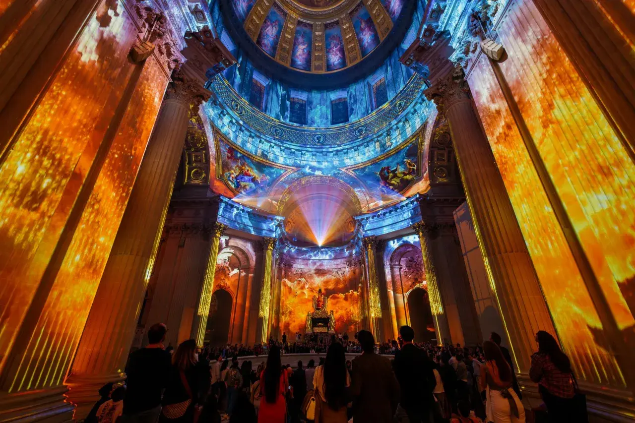 Spectacle lumineux projeté sur des colonnes et un dôme, créant une aura de couleurs vives. Une foule admire cette installation, comme si les dates invalides s'effaçaient.