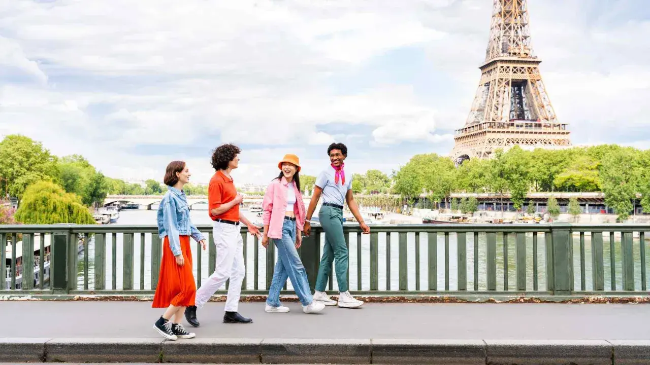 Quatre jeunes amis profitent d'une promenade ensoleillée à Paris, avec la Tour Eiffel en arrière-plan. L'un d'eux, un étudiant, porte un foulard rose vif.