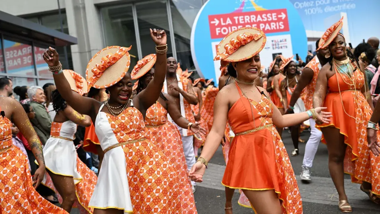 Des danseuses en tenues orange et blanches, chapeaux assortis, célèbrent la foire de Paris 2026 dans une ambiance festive.