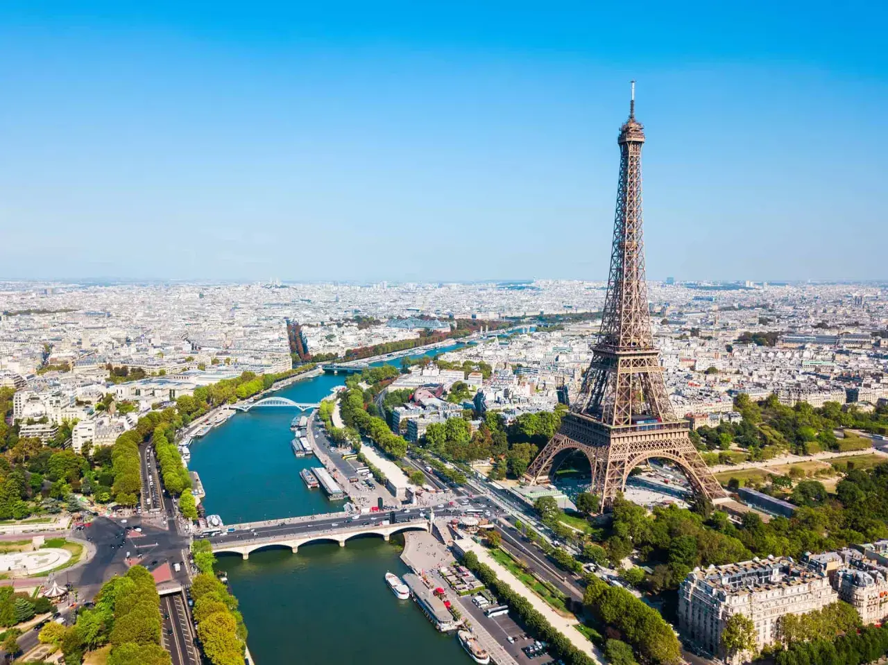 Vue aérienne de Paris avec la tour Eiffel dominant la ville. La Seine serpente à travers le paysage urbain, bordée d'arbres et de ponts.