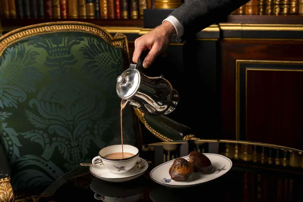 Un homme verse un chocolat chaud dans une tasse, accompagné de madeleines, dans un salon parisien luxueux.