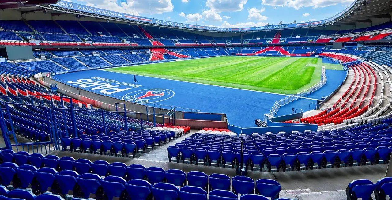 Vue panoramique du **Parc des Princes** vide, avec ses tribunes bleues et rouges, son terrain de football verdoyant et le logo du PSG au centre.