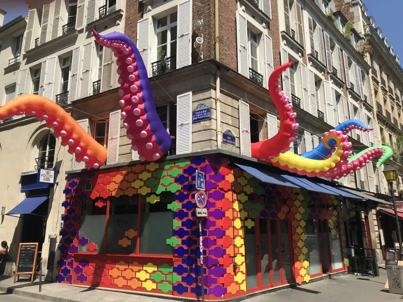 Tentacules colorés géants sortent d'un bâtiment orné de motifs arc-en-ciel, un décor festif pour ce bar gay à Paris.