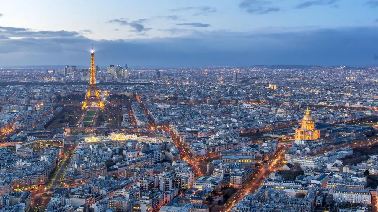 Vue panoramique de Paris au crépuscule, avec la Tour Eiffel illuminée et le dôme des Invalides. On aperçoit le quartier de La Défense au loin. Un panorama depuis l'**étage tour Montparnasse**.