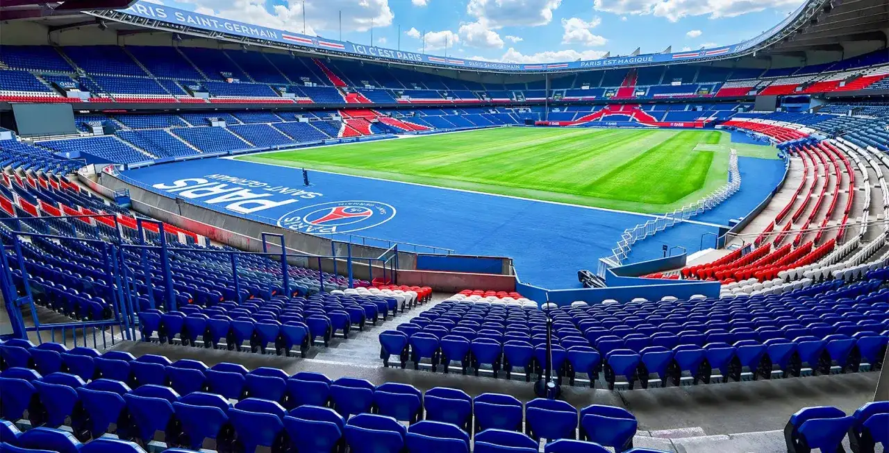 Vue panoramique du **Parc des Princes** vide, avec ses tribunes bleues et rouges, son terrain de football verdoyant et le logo du PSG au centre.