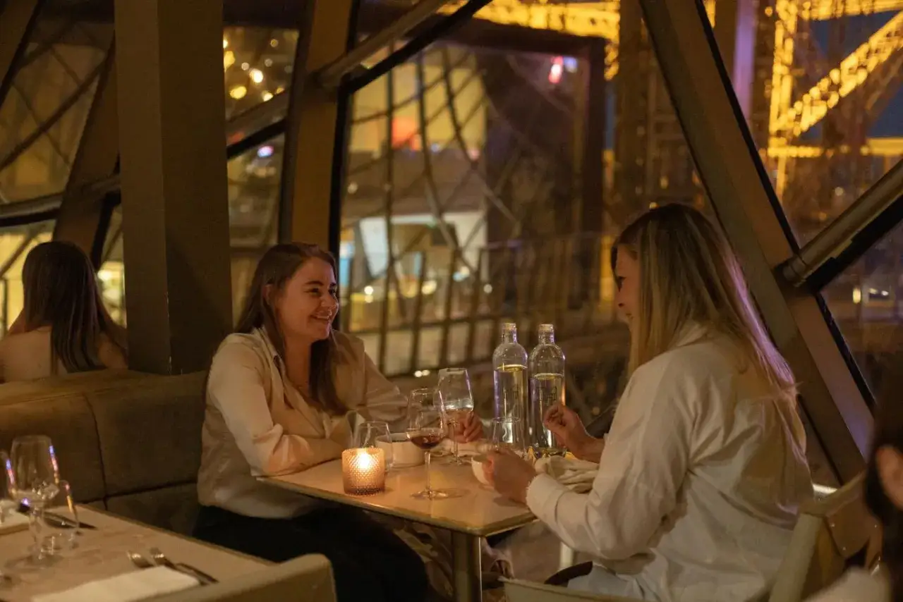 Deux femmes dînent dans une brasserie avec la Tour Eiffel illuminée en arrière-plan.