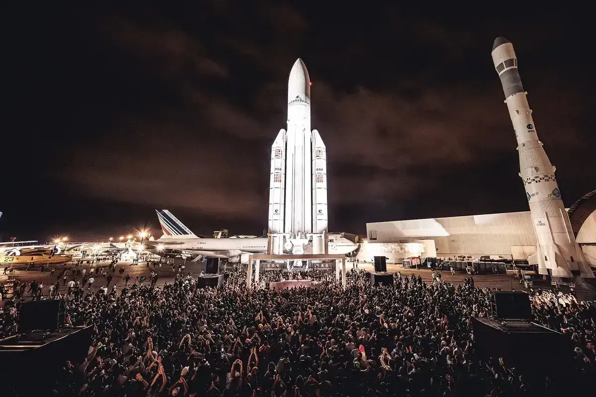 Foule immense devant une fusée et un avion, ambiance de cercle festival nocturne.