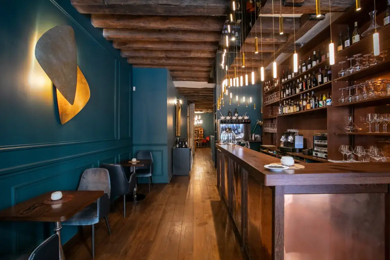 Ambiance chaleureuse d'un bar &agrave; vin parisien avec ses poutres apparentes, son mur bleu canard et son comptoir en bois.