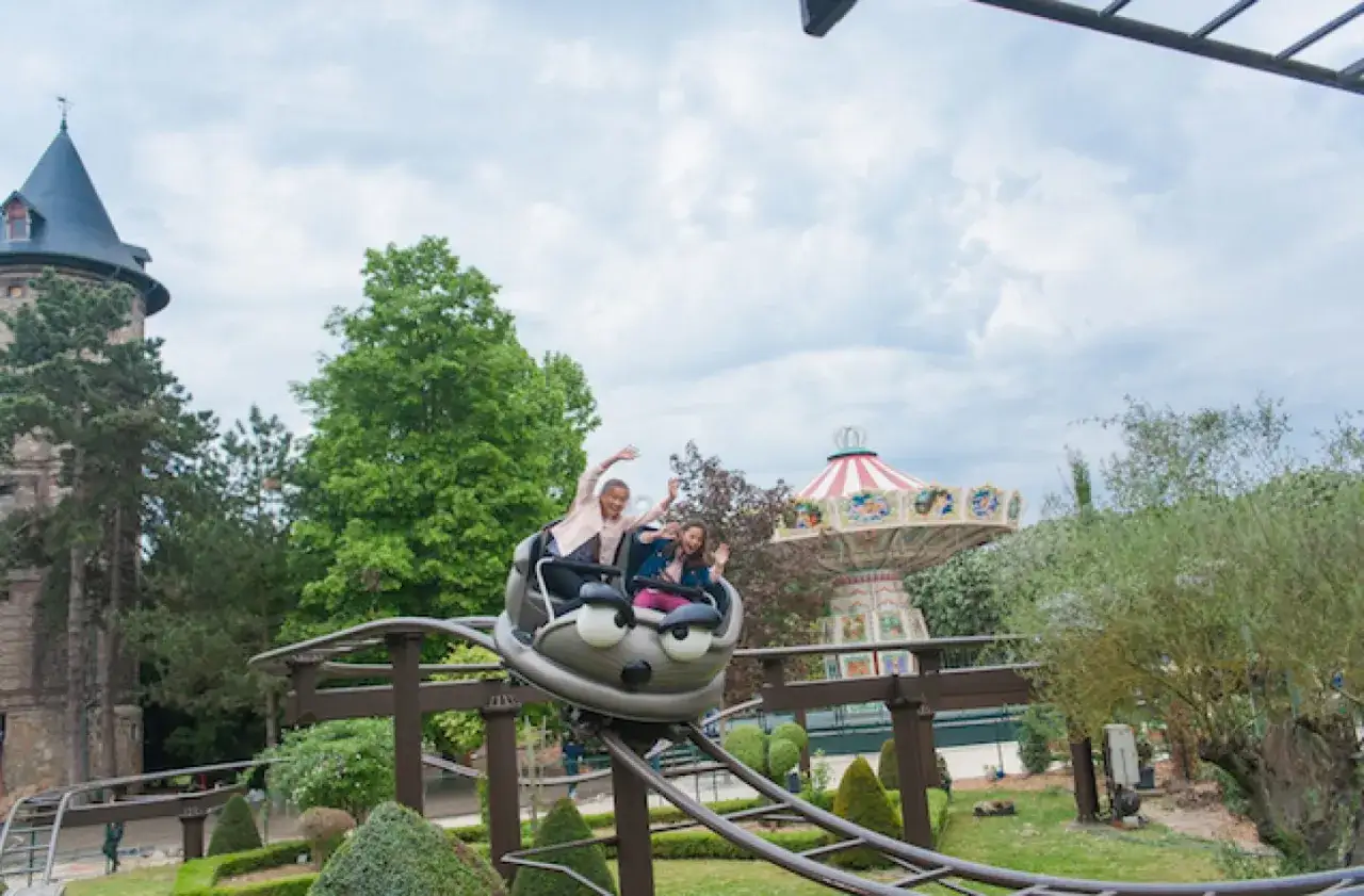 Deux enfants s'amusent dans un wagon de montagnes russes en forme de chenille dans un parc d'attractions. Un carrousel et un ch&acirc;teau compl&egrave;tent le d&eacute;cor.