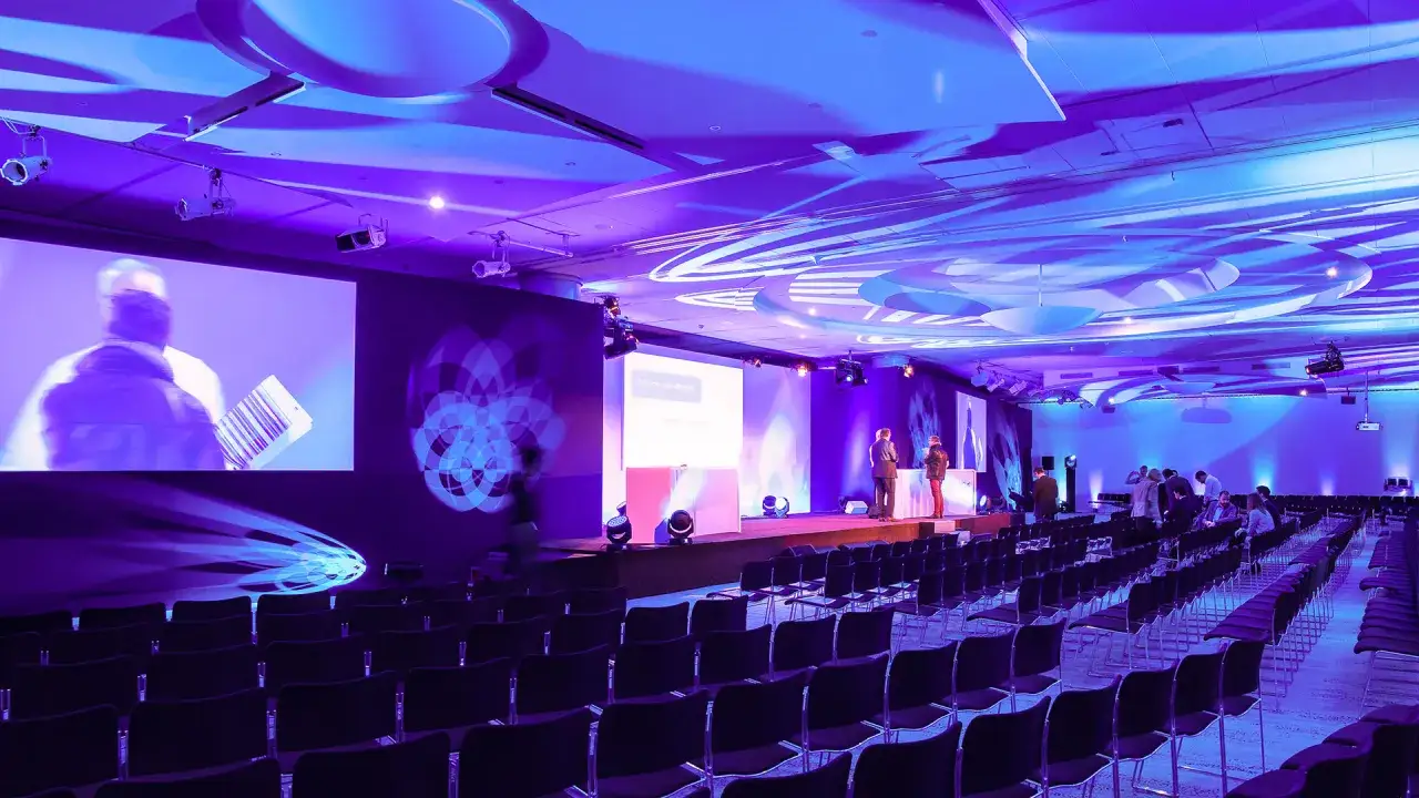 Salle de conf&eacute;rence illumin&eacute;e en violet, avec des rang&eacute;es de si&egrave;ges vides. Le nombre de places est impressionnant pour cet &eacute;v&eacute;nement de maroquinerie.
