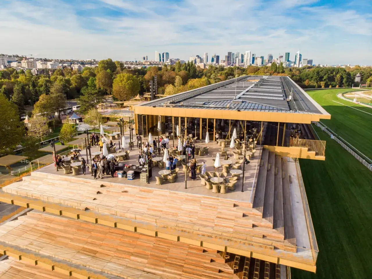 Vue a&eacute;rienne du toit d'un b&acirc;timent moderne avec des tables et des parasols sur une terrasse, avec le paysage urbain de Paris et l'hippodrome de Longchamp en arri&egrave;re-plan.