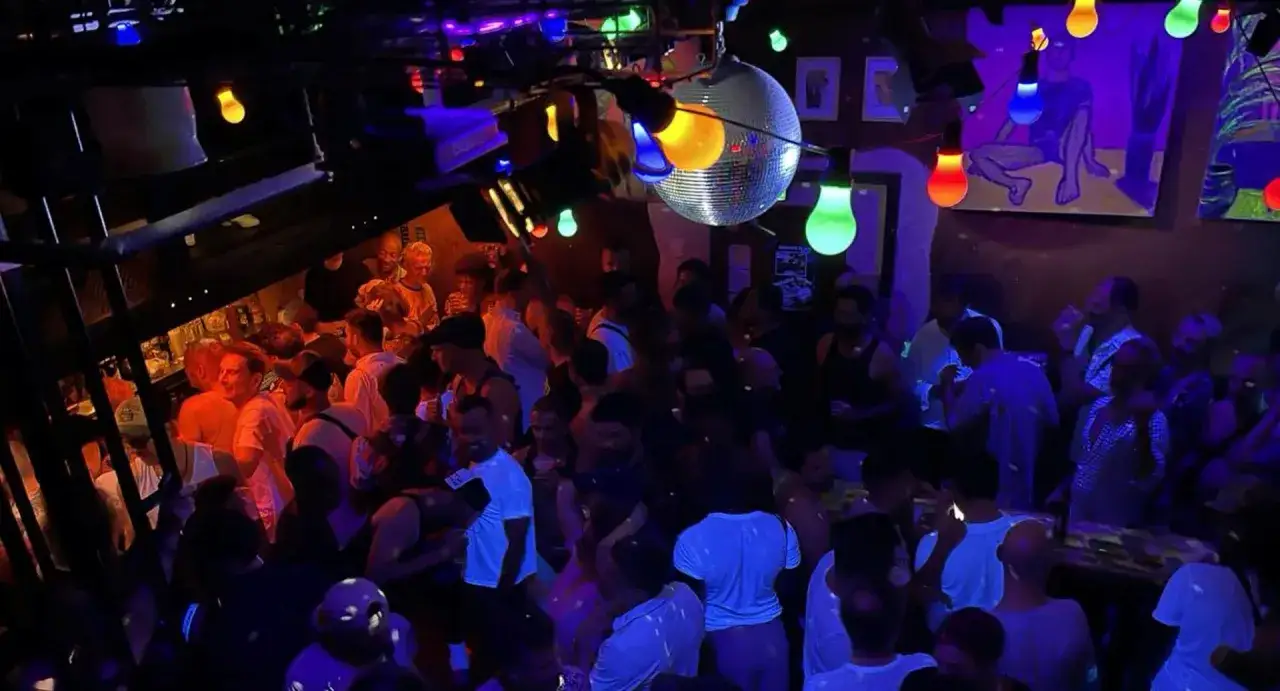 Ambiance festive dans un bar gay, avec une foule anim&eacute;e sous des lumi&egrave;res color&eacute;es et une boule disco.