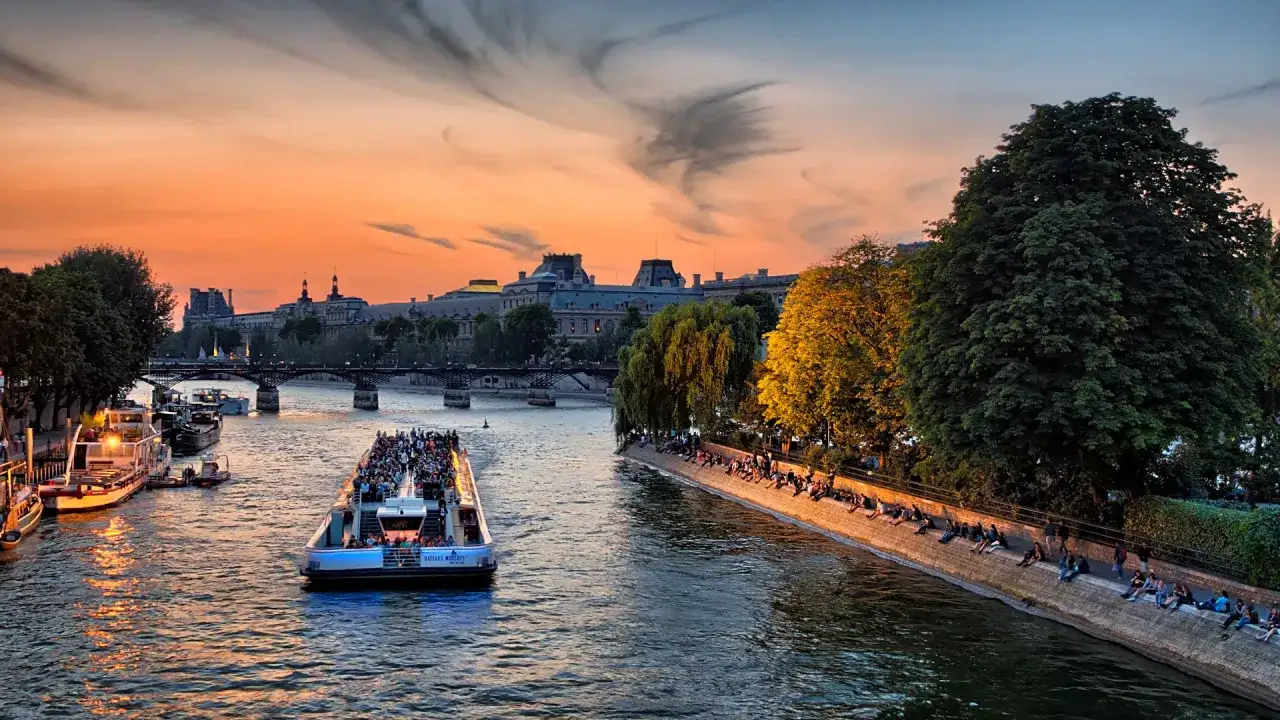 Coucher de soleil sur la Seine, Paris. Un bateau plein de touristes navigue, tandis que des gens profitent de la vue depuis les quais. Le ciel, un spectacle de couleurs, rappelle le talent d'un peintre, un vrai mazette.