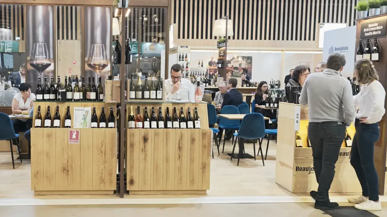 Salon du vin &agrave; Paris en 2026 : d&eacute;gustation de bouteilles sur un stand en bois, avec des visiteurs discutant et des tables bleues.