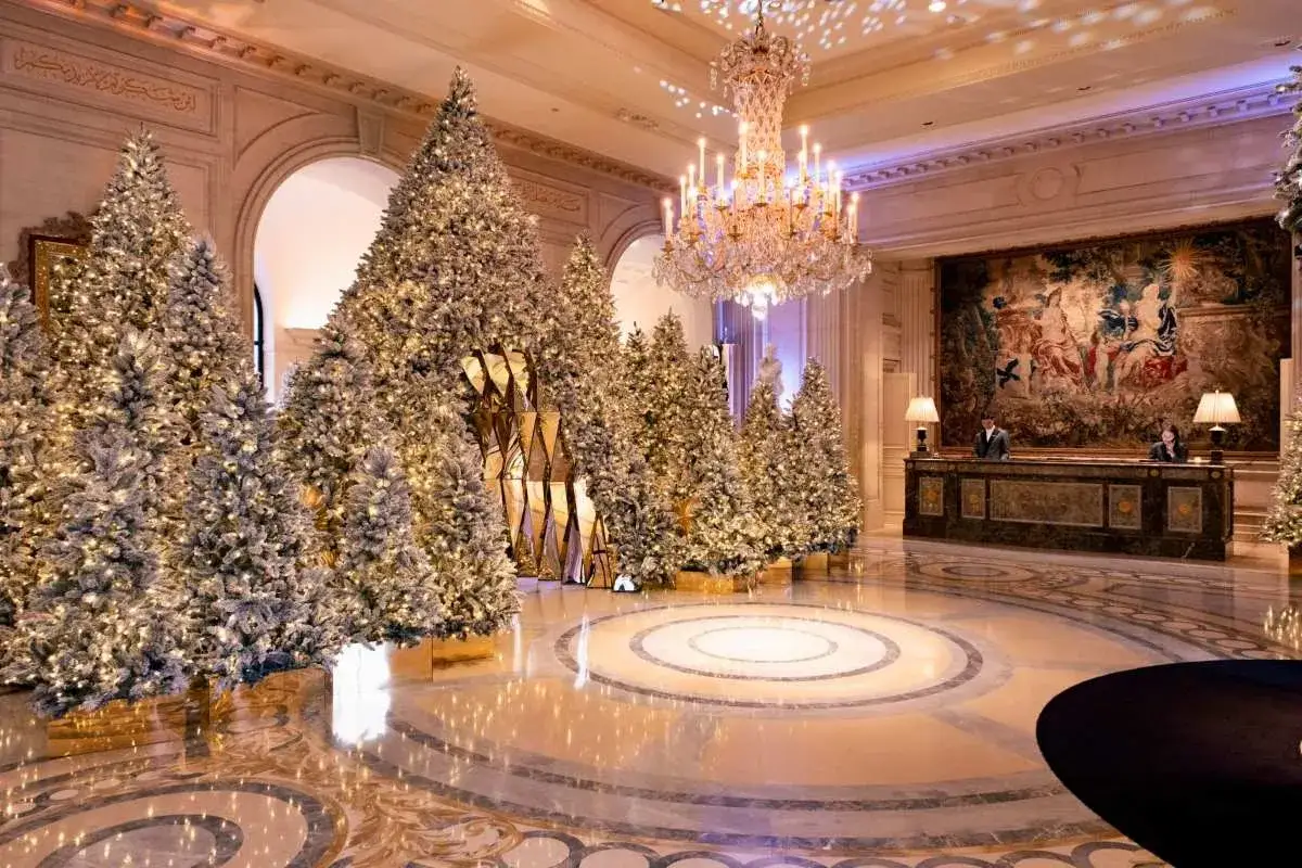 Un hall d'hôtel somptueux, décoré de nombreux sapins illuminés pour une soirée de Noël féerique. Un lustre scintille au plafond.