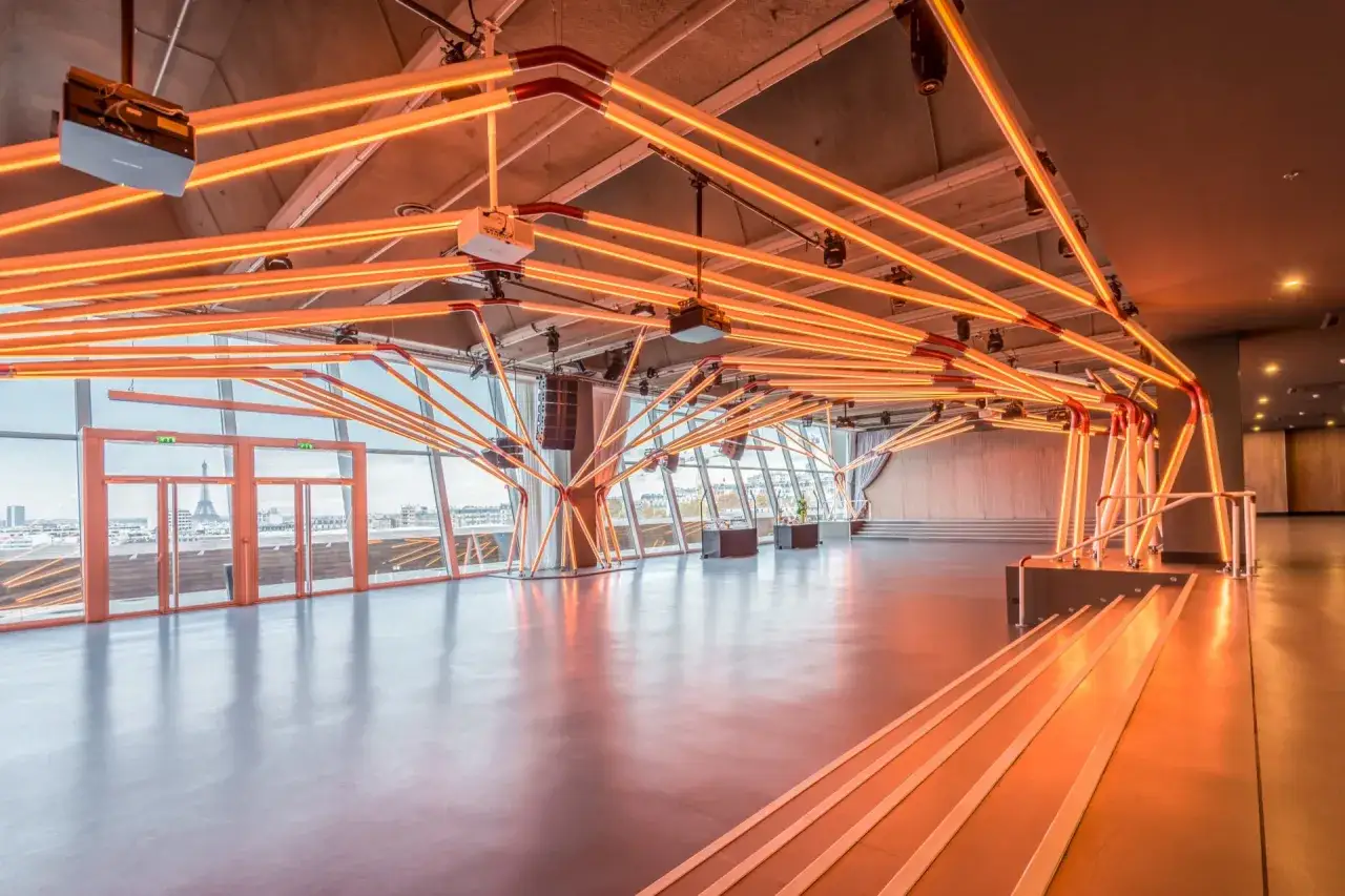 Un espace &eacute;v&eacute;nementiel moderne &agrave; Paris, illumin&eacute; par des n&eacute;ons orange vifs. La Tour Eiffel est visible &agrave; travers les grandes baies vitr&eacute;es.