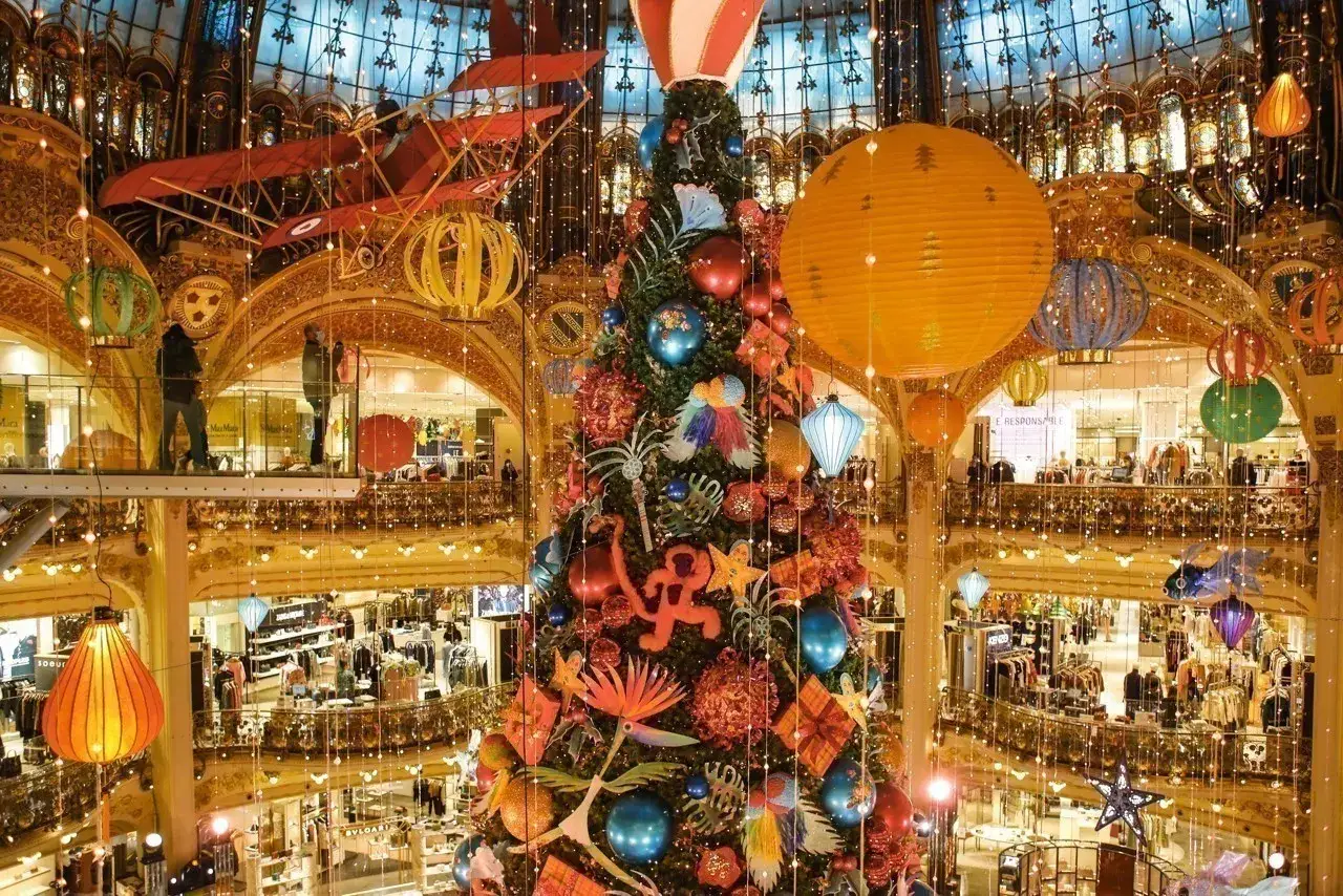 Un sapin de Noël majestueux au centre, décoré de boules colorées et de guirlandes lumineuses. Au-dessus, un avion rouge et des lanternes flottent, créant une ambiance féerique typique de la deco noel paris.