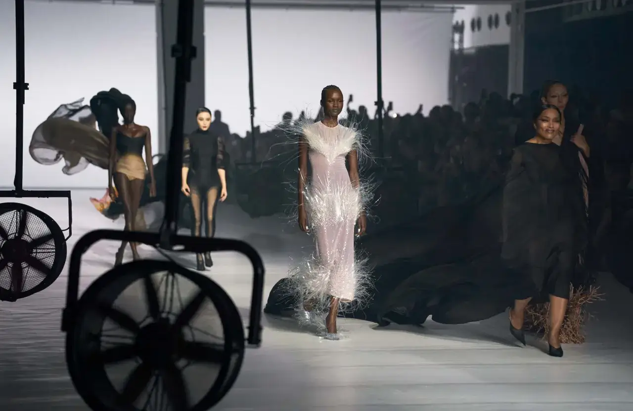 Mannequins défilent sur un podium, une robe ornée de plumes blanches attire le regard. L'ambiance évoque la mode parisienne de 2026.