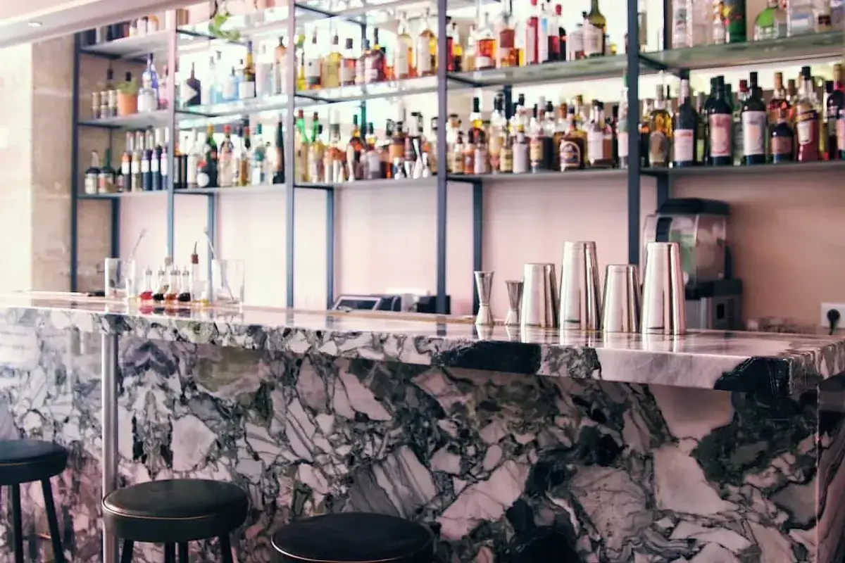Un bar élégant avec un comptoir en marbre veiné, des tabourets noirs et des étagères remplies de bouteilles. Le menu de bar-bisou promet des délices.