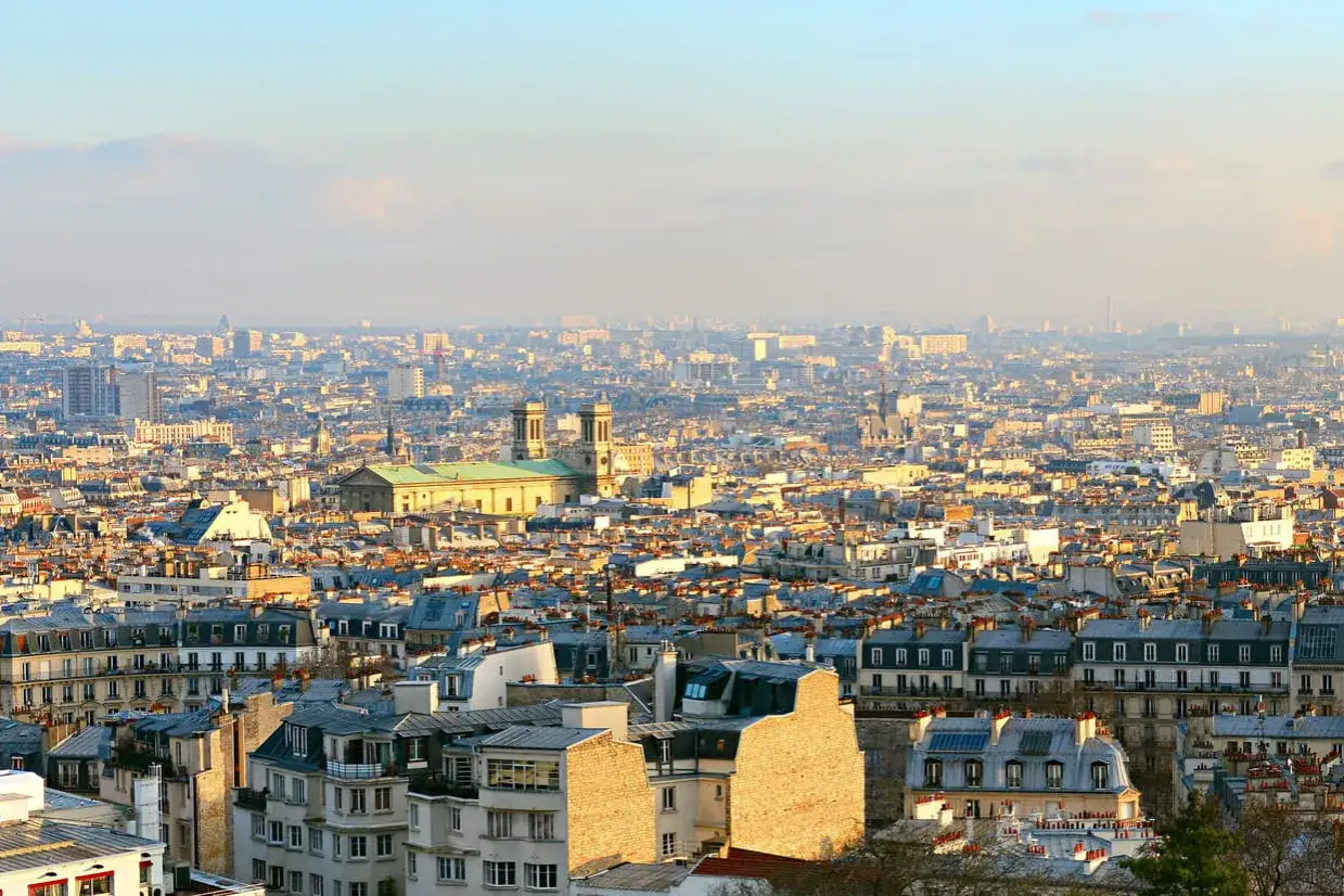 Vue panoramique de Paris, avec le Sacré-Cœur dominant la ville. Les toits de zinc et les façades haussmanniennes s'étendent à perte de vue sous un ciel clair.