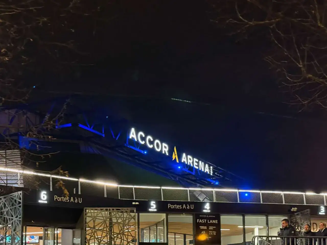 La nuit tombe sur l'Accor Arena, illumin&eacute;e de n&eacute;ons bleus. Les portes d'entr&eacute;e sont visibles, pr&ecirc;tes &agrave; accueillir les spectateurs.