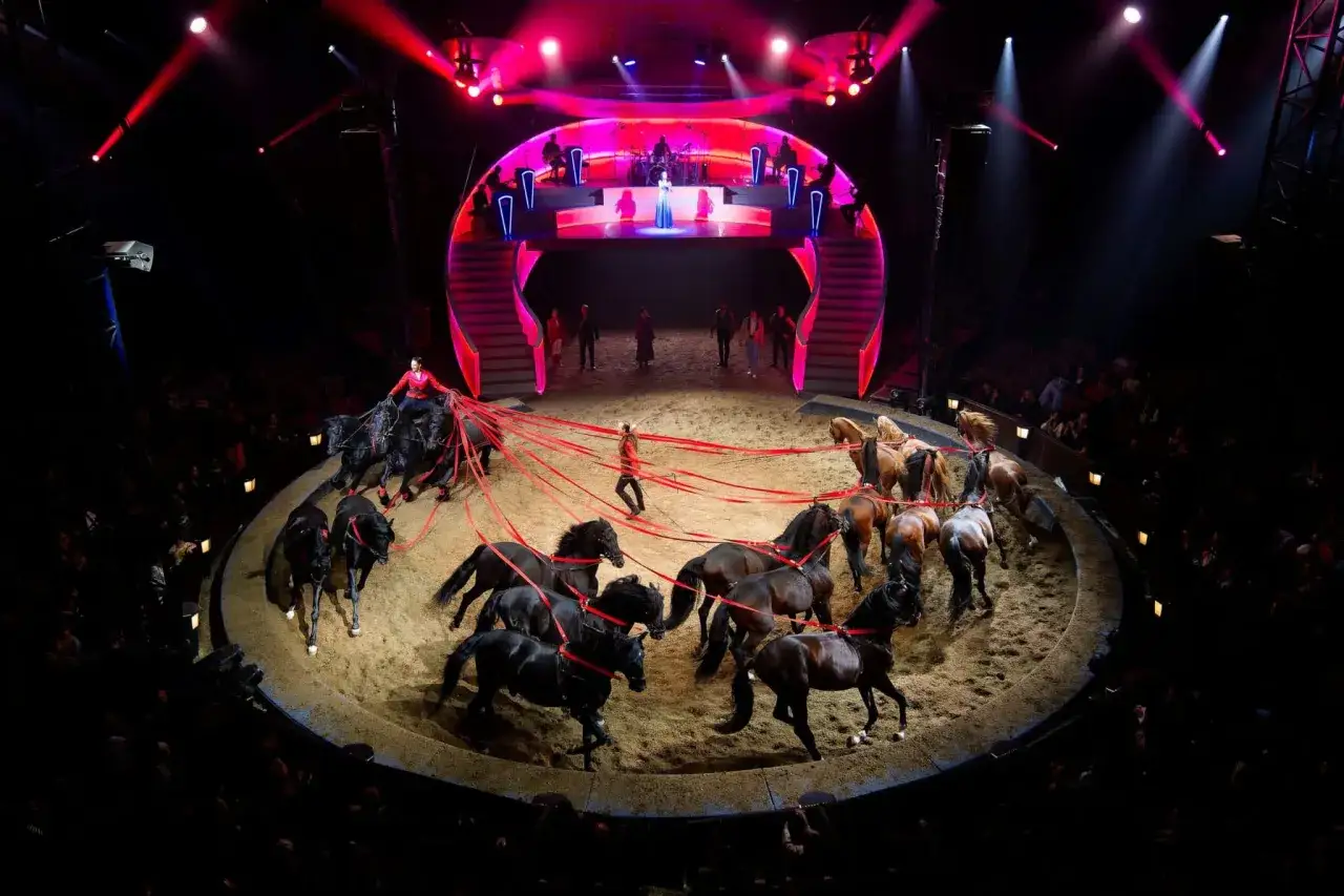 Un dresseur dirige une troupe de chevaux noirs et bruns dans le cirque des Folies Gruss, sous des lumières rouges vives.
