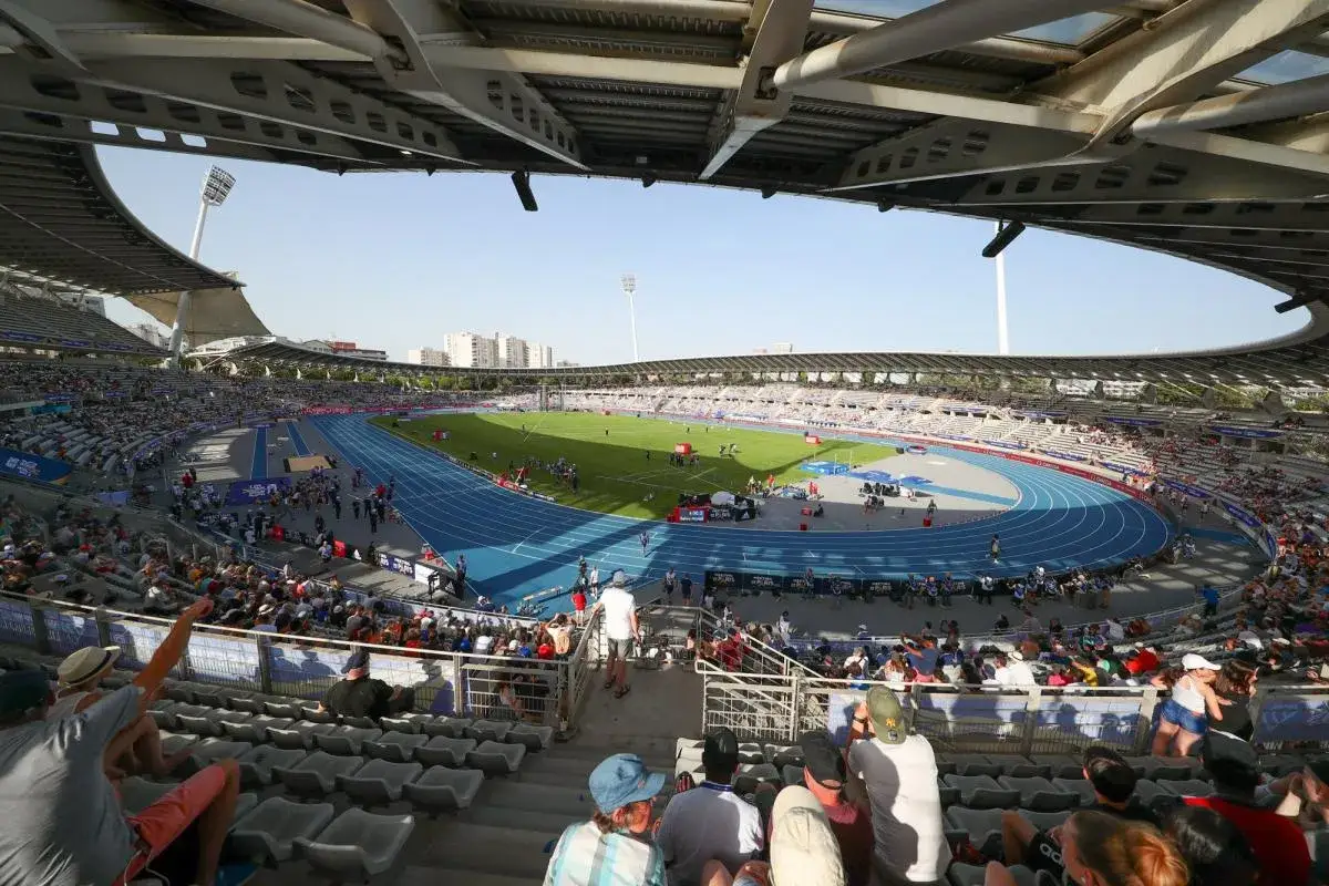 Vue panoramique du stade Charl&eacute;ty, avec une piste d'athl&eacute;tisme bleue, un terrain de football vert et des gradins remplis de spectateurs sous un ciel bleu.
