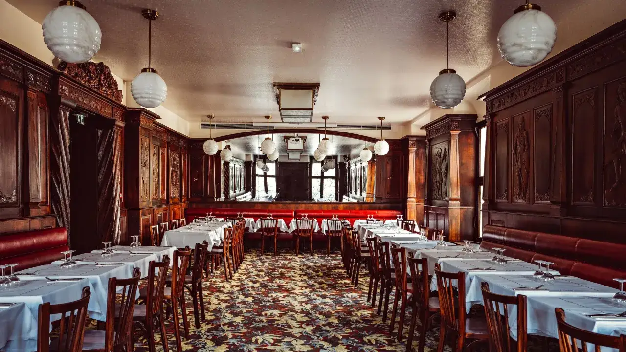 Salle de restaurant avec tables dressées, banquettes rouges et boiseries sculptées. Ambiance feutrée digne d'un bouillon république.