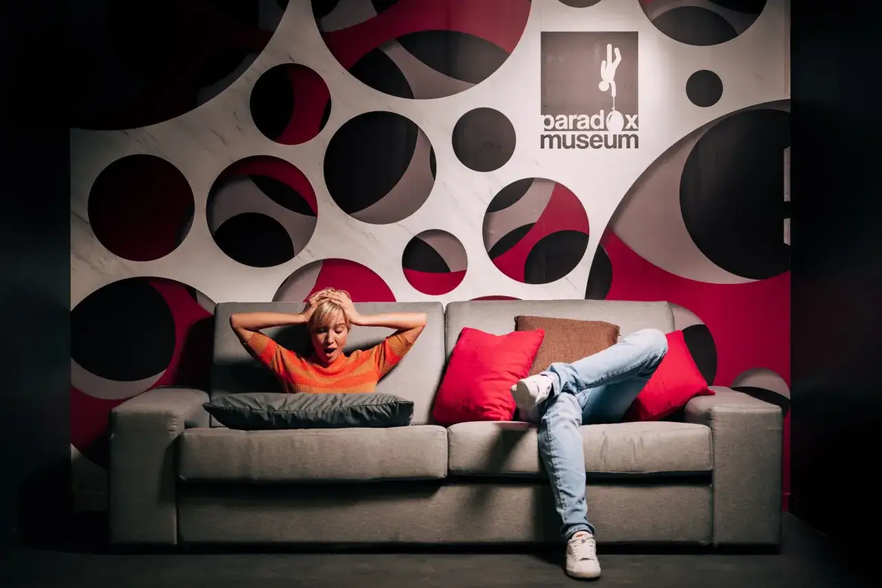Une femme choqu&eacute;e sur un canap&eacute; devant un mur de cercles. Le logo du Paradox Museum est visible. La dur&eacute;e de visite du mus&eacute;e de l'illusion est une exp&eacute;rience inoubliable.