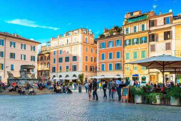 Une place animée à Rome, avec des bâtiments colorés et une fontaine. Un panneau indique "Take Away Cocktail 4.50", un clin d'œil au "saint amour menu".
