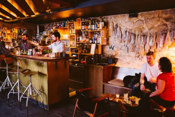 Ambiance feutrée dans ce bar caché. Un barman sert un cocktail, des clients discutent au comptoir et sur une banquette.