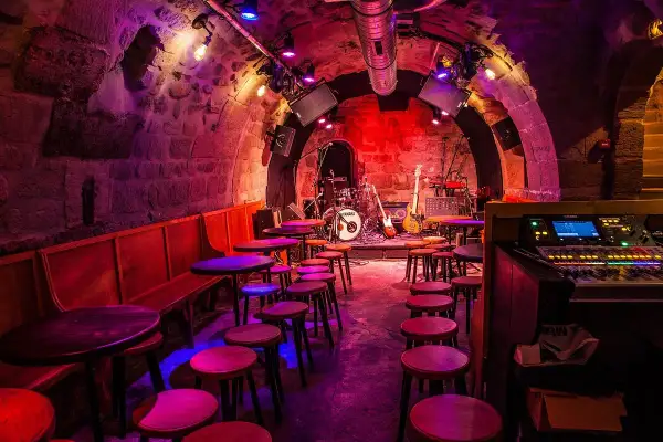 Scène de concert dans le caveau des oubliettes, éclairée par des lumières violettes et rouges. Tables et tabourets disposés devant la scène où se trouve une batterie et des guitares.
