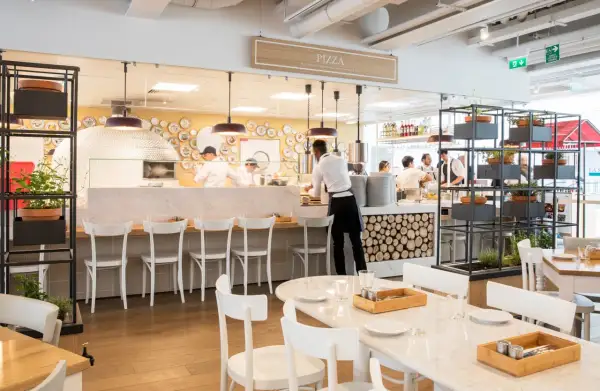 Un restaurant italien moderne avec un four à pizza visible. Des chefs préparent des plats derrière le comptoir, tandis que des tables sont dressées pour les clients.