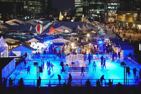 Patinage sur glace et marchés de Noël ouverts, illuminés par des lumières bleues vives. Une scène festive avec des chalets et des décorations scintillantes.
