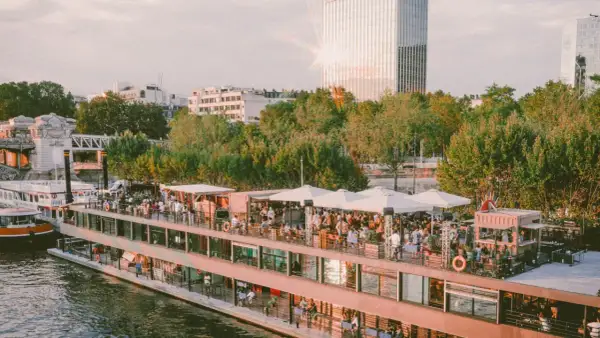 Un bateau-mouche animé sur la Seine, avec des gens faisant la fête sur le pont. Le soleil brille sur un gratte-ciel, créant un effet de "mazette" lumineux.