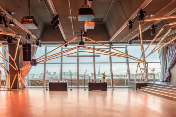 Salle de réception moderne avec vue panoramique sur Paris, éclairage néon, bar et scène. La Tour Eiffel est visible au loin.