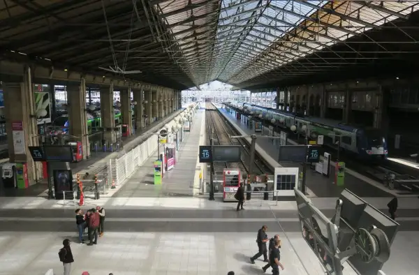 Intérieur lumineux de la gare du Nord, avec des quais, des voies ferrées et des trains. Des voyageurs circulent sous la grande verrière.