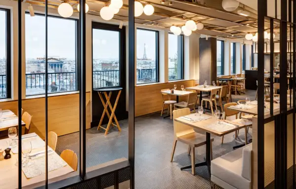 Restaurant moderne avec vue sur Paris. Tables dressées, lumières douces, et le logo "Printemps du Goût" visible.
