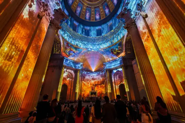 Spectacle lumineux projeté sur des colonnes et un dôme, créant une aura de couleurs vives. Une foule admire cette installation, comme si les dates invalides s'effaçaient.